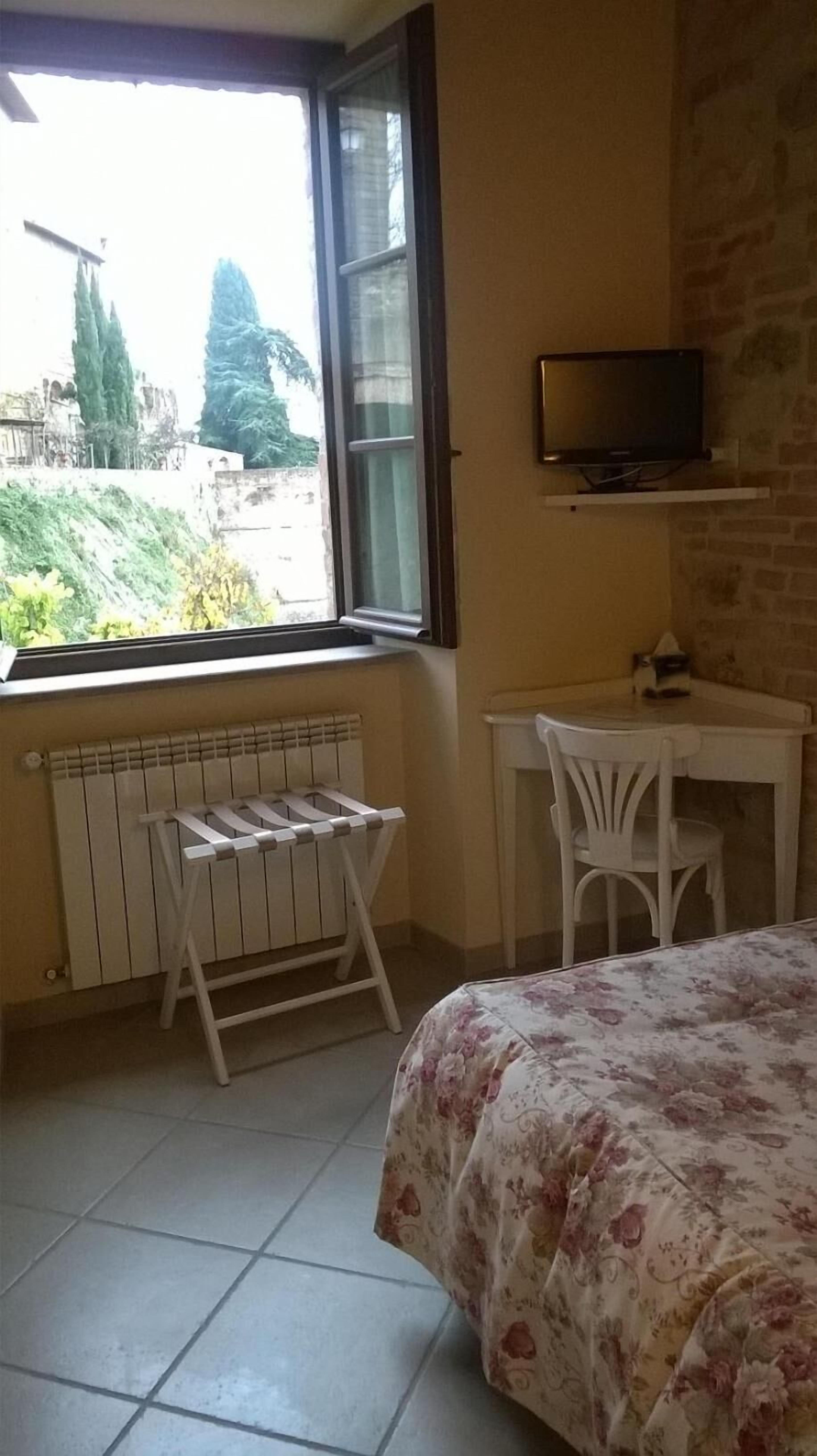 A bedroom with a bed, a desk, a chair, a radiator, and a window with a view of trees and buildings.