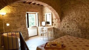 A rustic room with a stone wall, a wooden desk, a chair, a bed, and a small table with food. - Arnolfo B&B (Colle di Val d'Elsa)