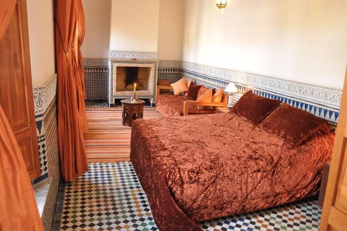 A room with a large bed, a small table with a lamp, and a fireplace.