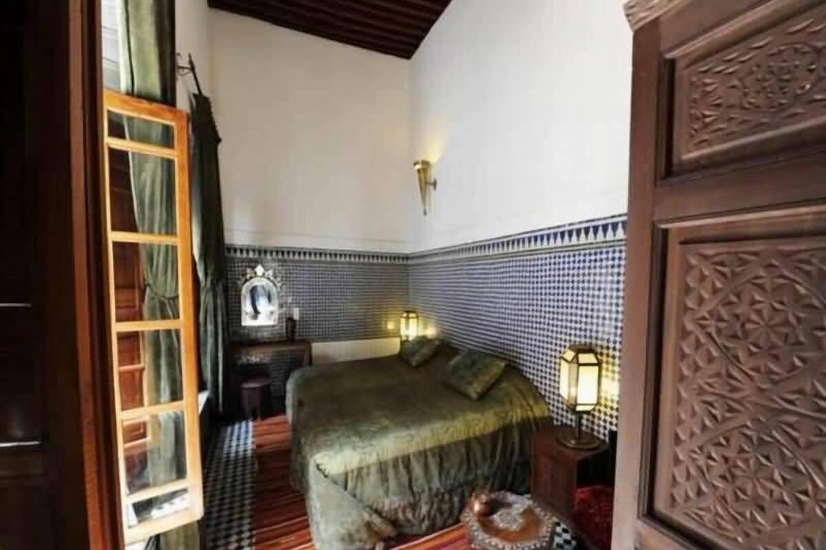 A bedroom with a checkered floor, a bed with a green bedspread, two bedside lamps, and a decorative wall panel.