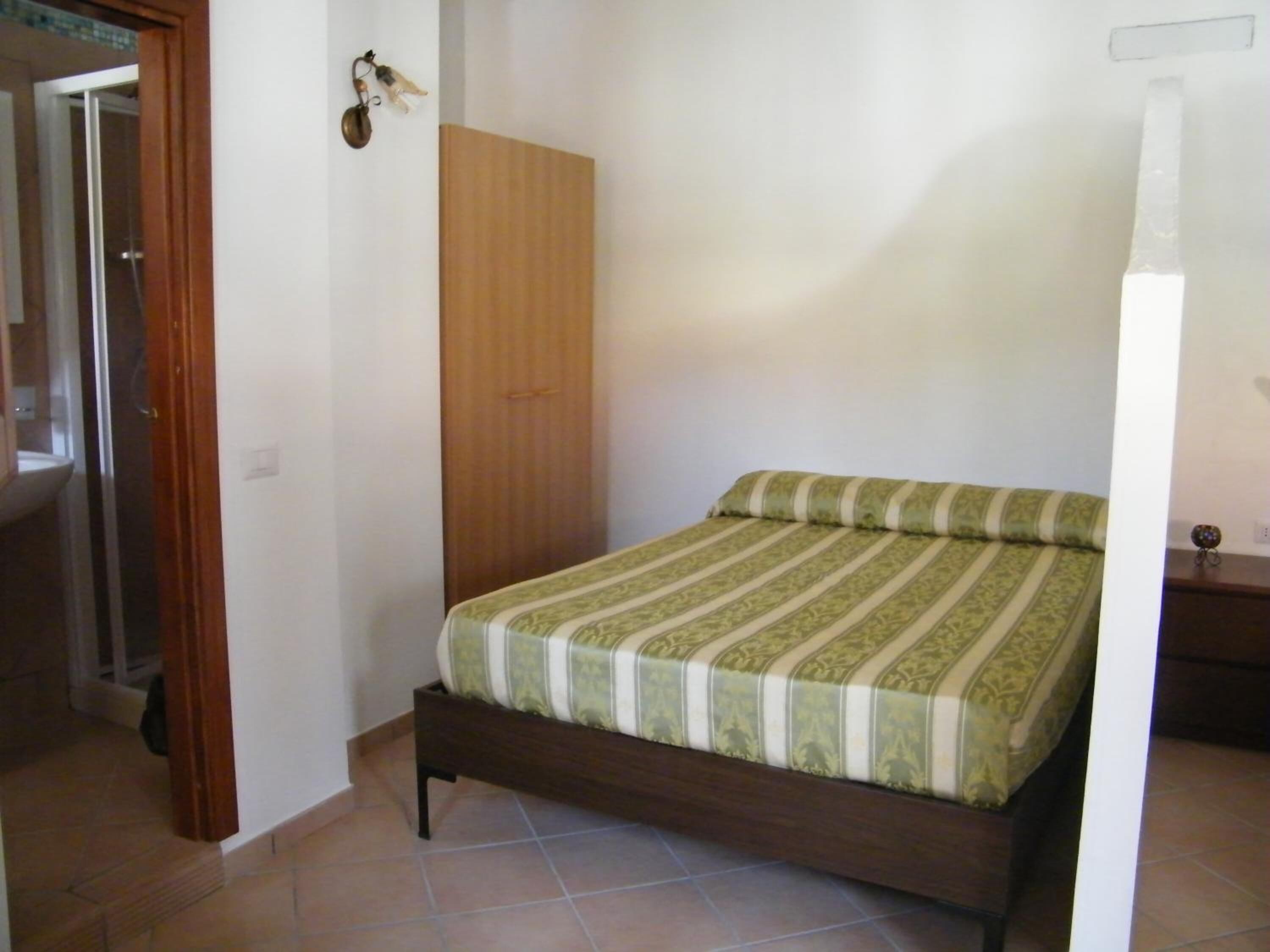 A bedroom with a bed, a wooden wardrobe, and a bathroom visible through an open door.