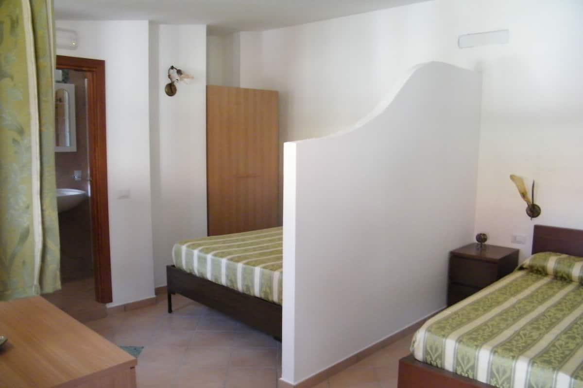 A bedroom with a bed, a nightstand, a wooden wardrobe, and a bathroom visible through an open door.