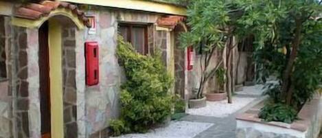 Un edificio de piedra con techo de tejas, un camino que lleva a una puerta y un pequeño jardín con árboles y arbustos.