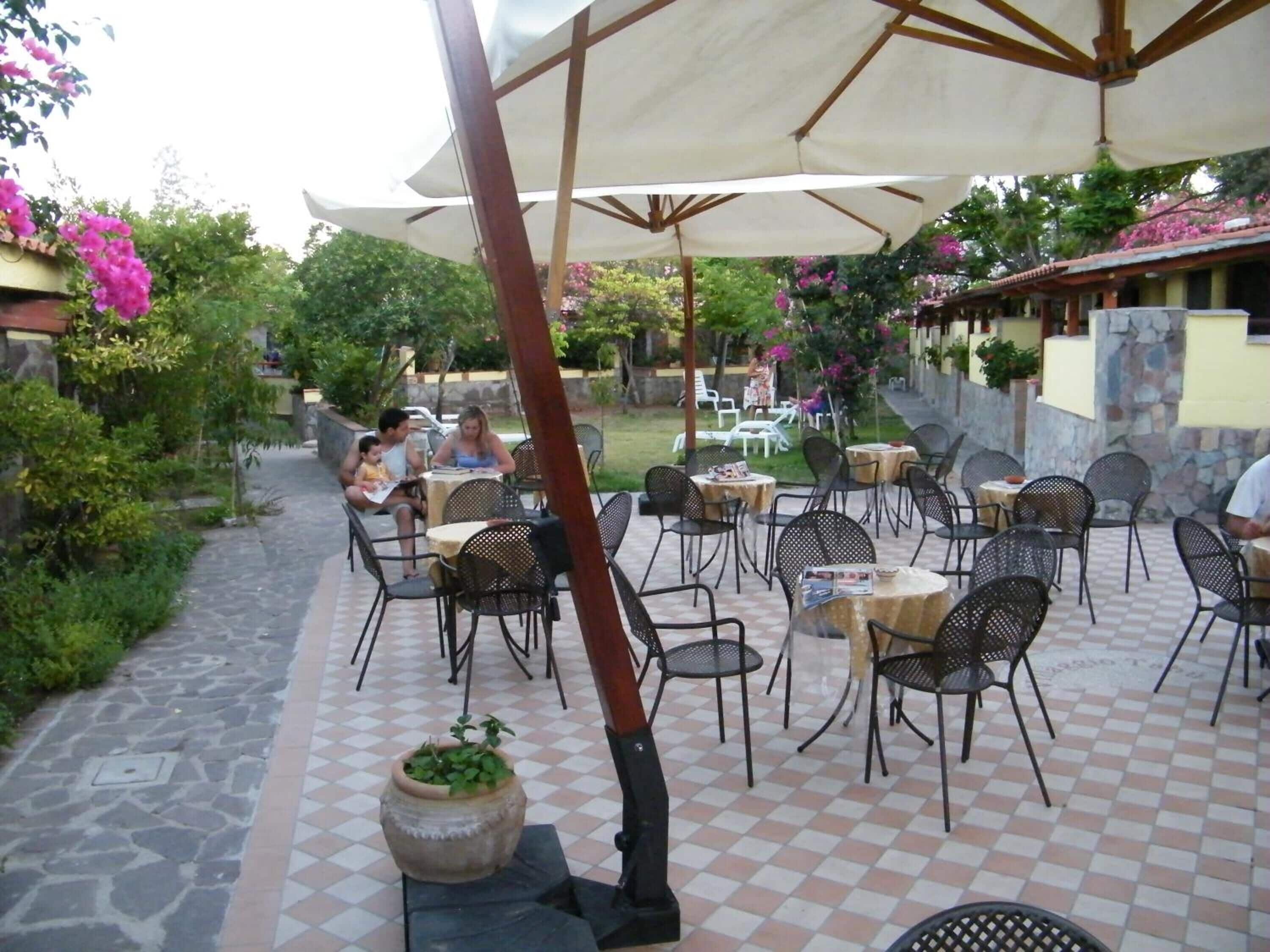An outdoor patio with tables and chairs, surrounded by greenery and a stone wall.