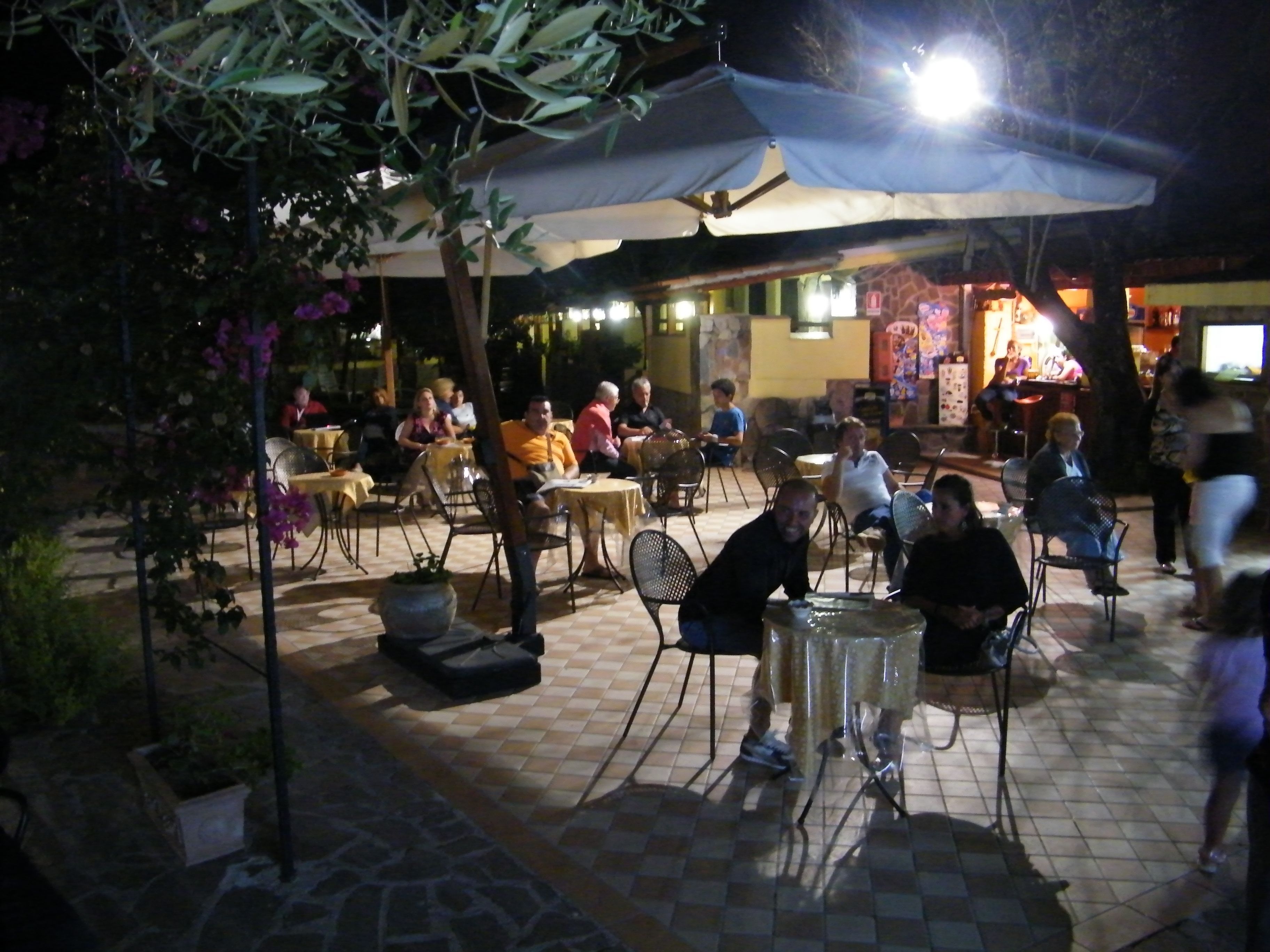 An outdoor café with tables and chairs under umbrellas, surrounded by potted plants and a stone floor.