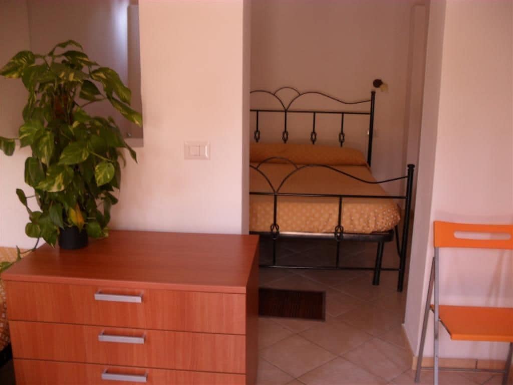 Ein Schlafzimmer mit einem Bett, einer Holzkommode, einer Zimmerpflanze und einem Sessel.