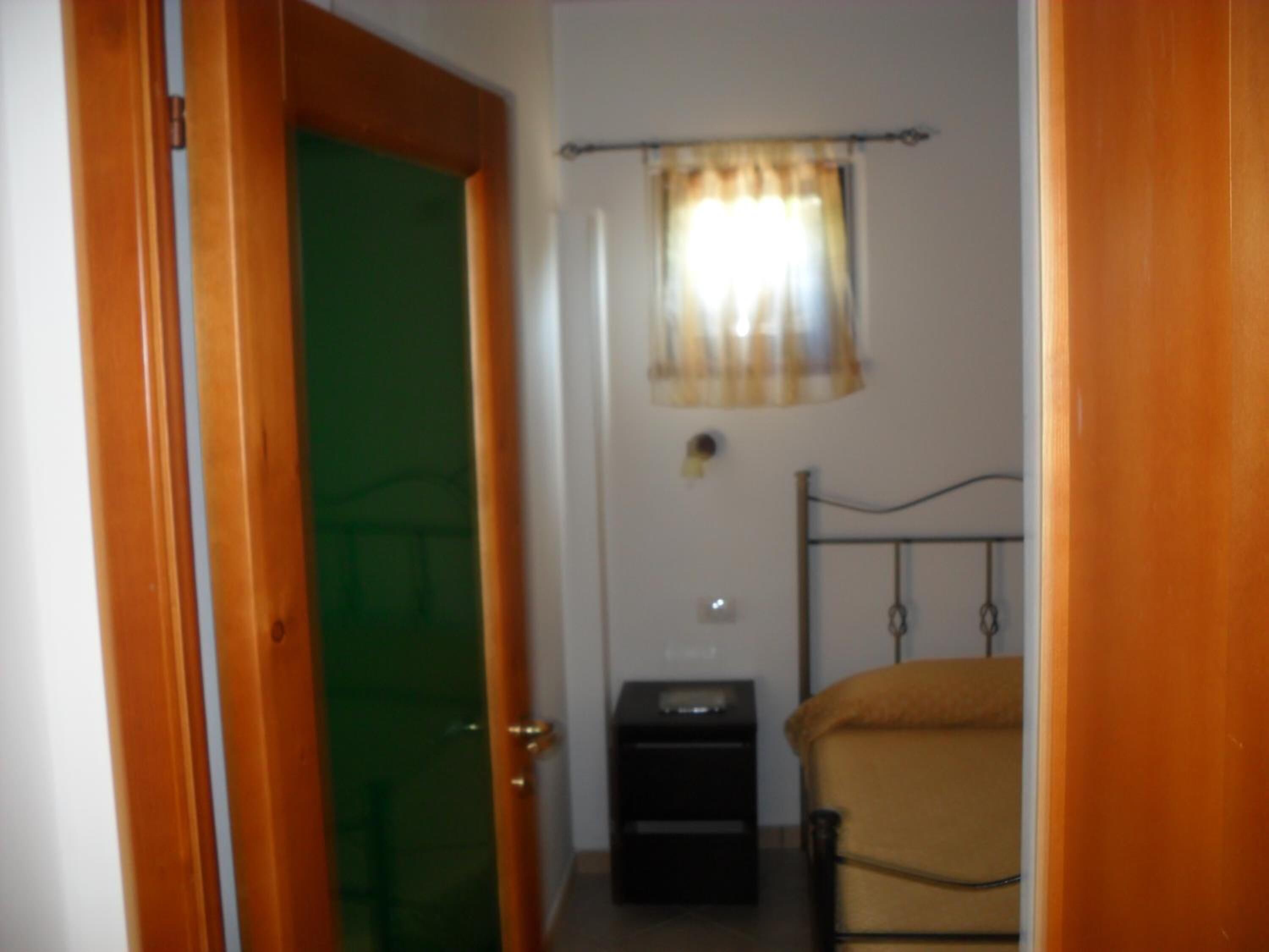 A small room with a wooden door, a green door frame, a small bedside table, a bed with a yellow blanket, and a window with a curtain.