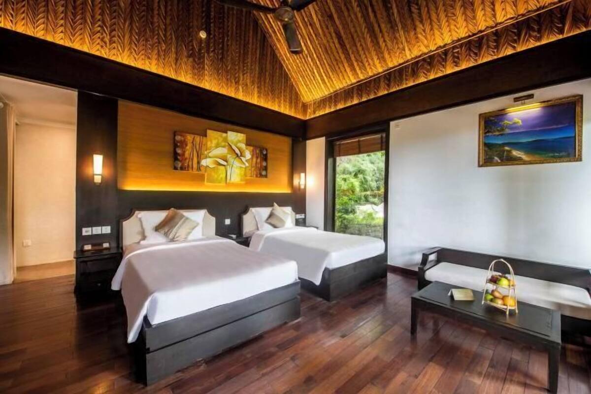 A hotel room with two beds, a sofa, a coffee table, a painting, and a view of greenery.