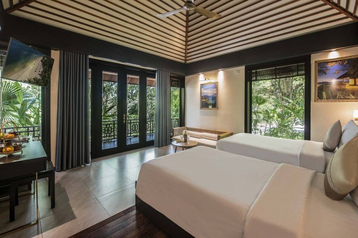 A modern hotel room with a large bed, a desk, and a balcony with a view of greenery.