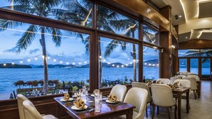 A restaurant with tables set for dining, large windows offering a view of the sea and palm trees, and string lights. - Hon Tam Resort (Nha Trang)
