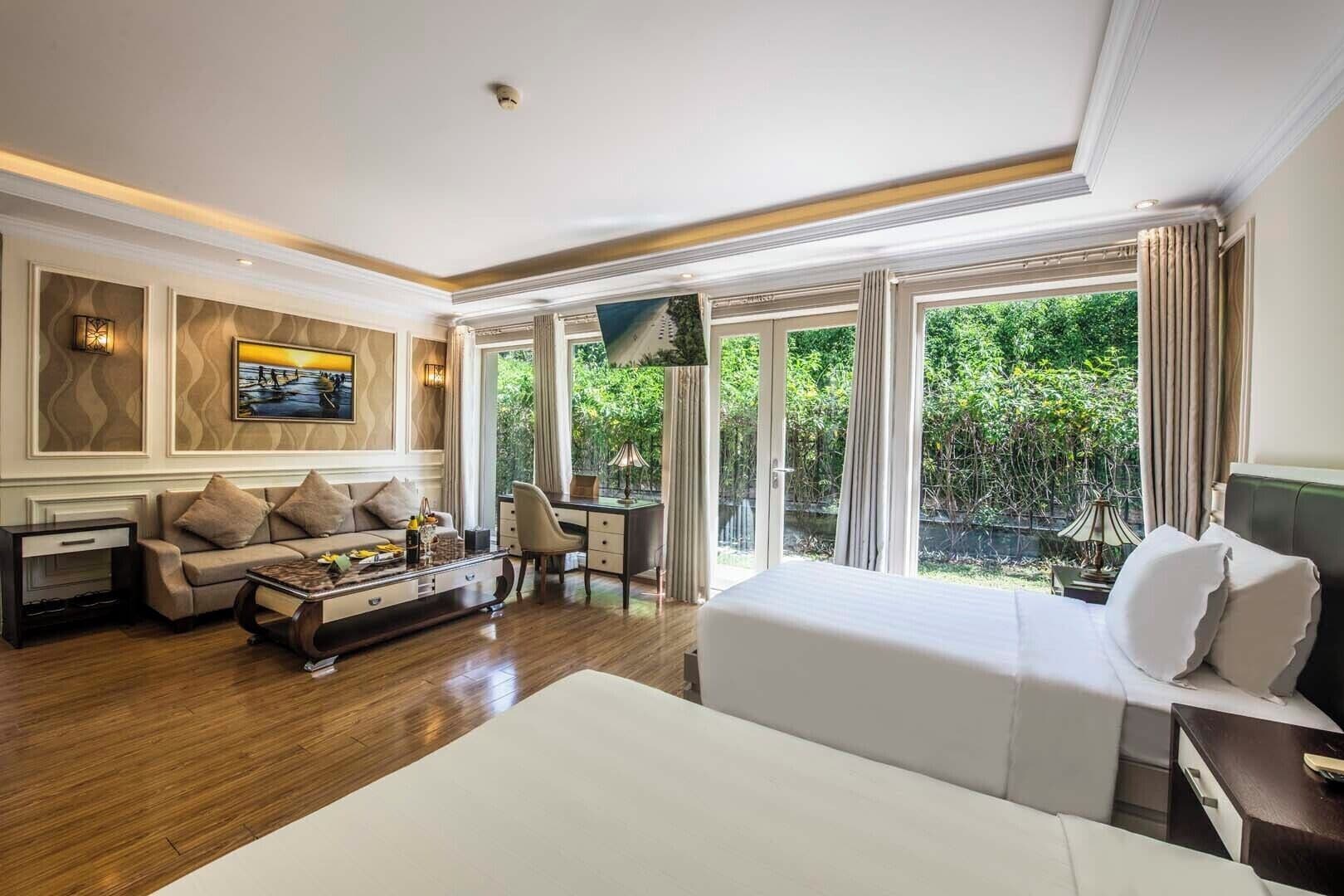A hotel room with a large bed, a sofa, a desk, and a view of greenery through large windows.