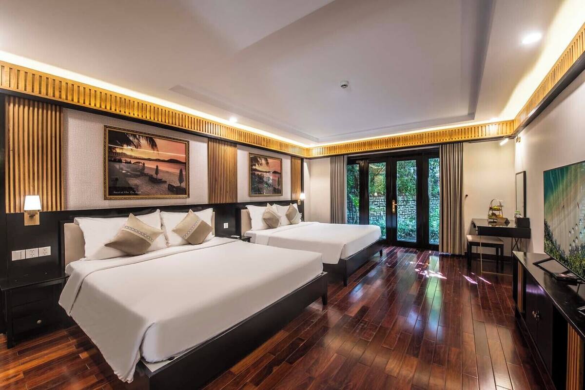 A hotel room with two beds, a desk, and a view of greenery through the door.