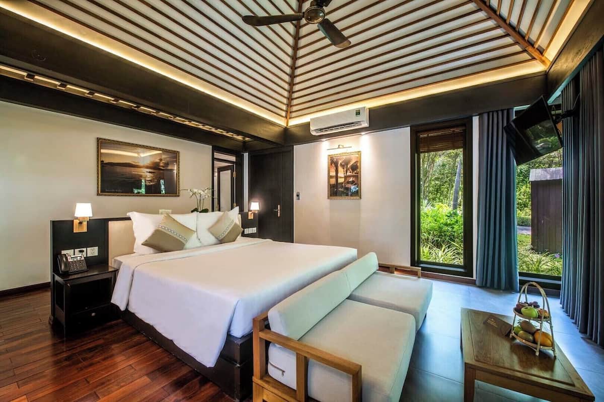 A hotel room with a large bed, bedside tables, a desk with a chair, a ceiling fan, and a large window with a view of greenery.