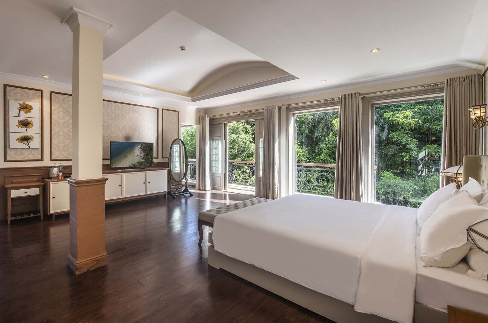 A spacious bedroom with a large bed, a vanity with a mirror, a television, and a balcony with a view of greenery.