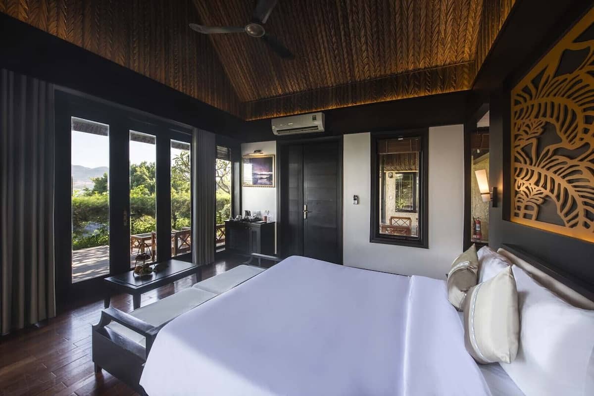 A bedroom with a large bed, a desk, and a chair, a window with a view of greenery, and a ceiling fan.