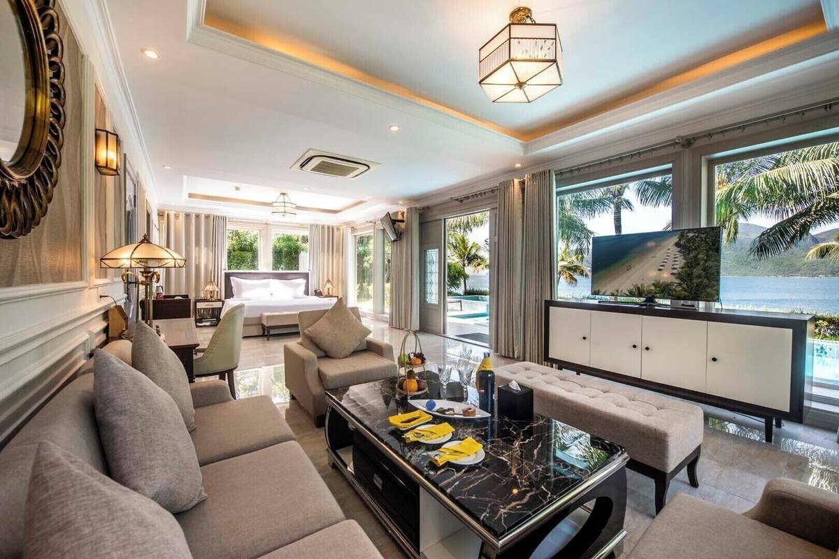 A spacious hotel room with a large bed, a sitting area with a sofa and armchairs, a dining table with a TV, and a view of the sea.