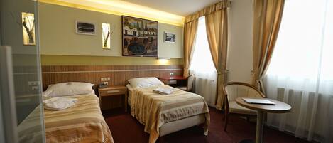 Habitación de hotel con dos camas individuales, un escritorio pequeño, una silla y una ventana con cortinas.