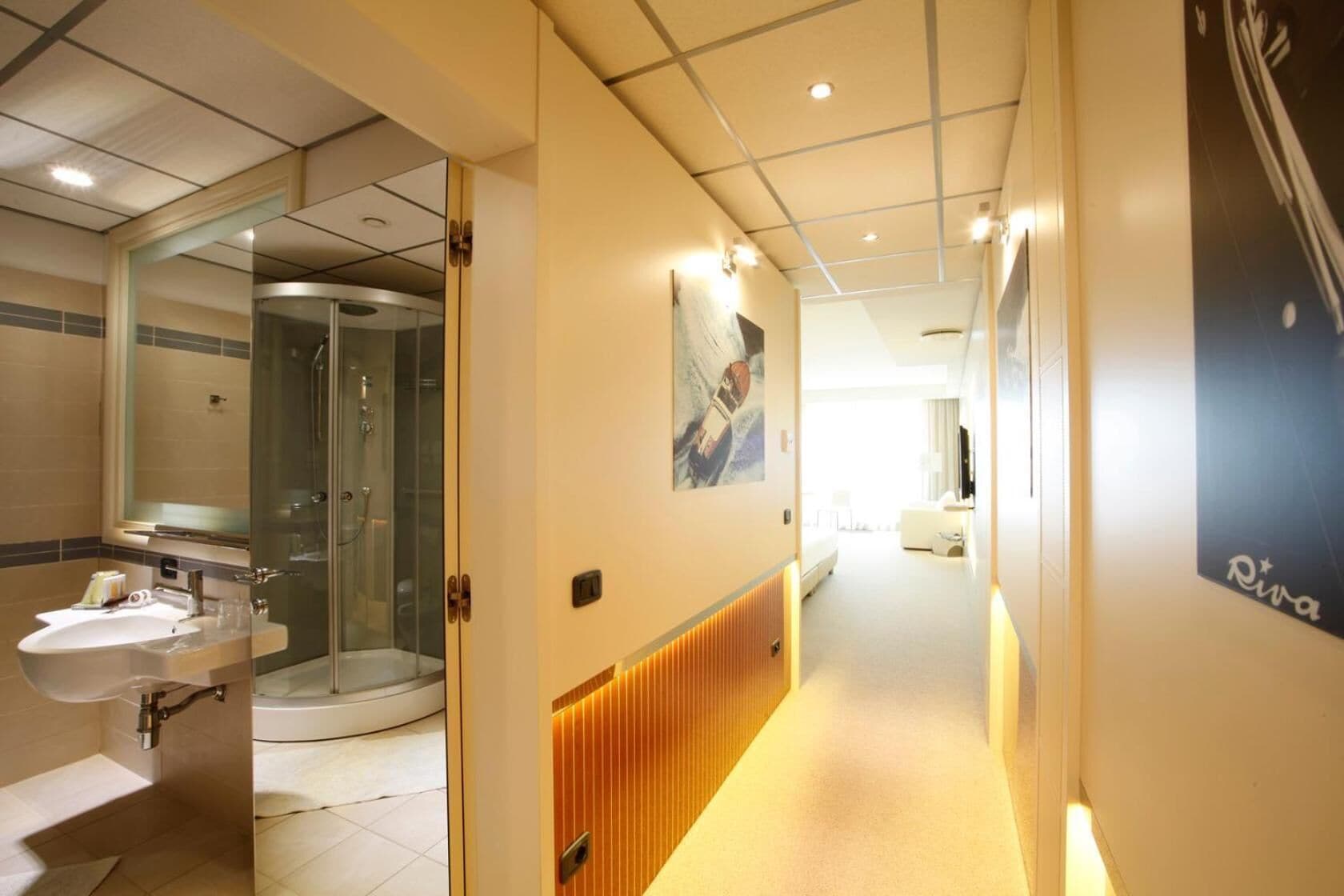 Un couloir d’hôtel avec une salle de bain à gauche, un mur recouvert de panneaux de bois et une affiche à droite.