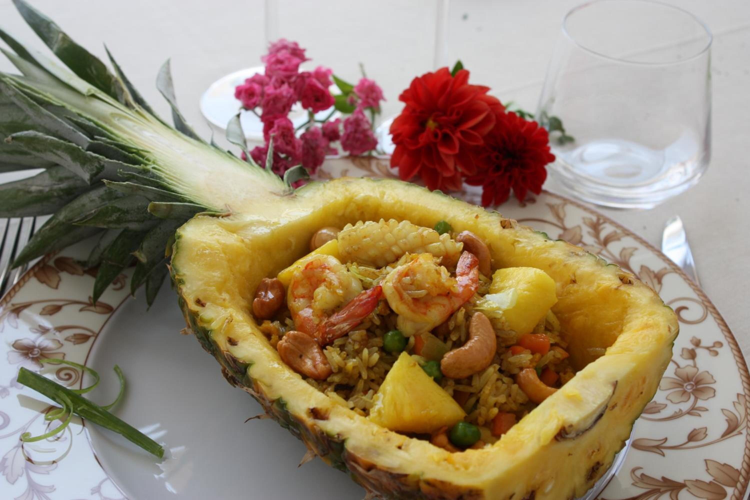 A plate of shrimp and rice dish in a pineapple shell, with flowers and a glass of water on the table.