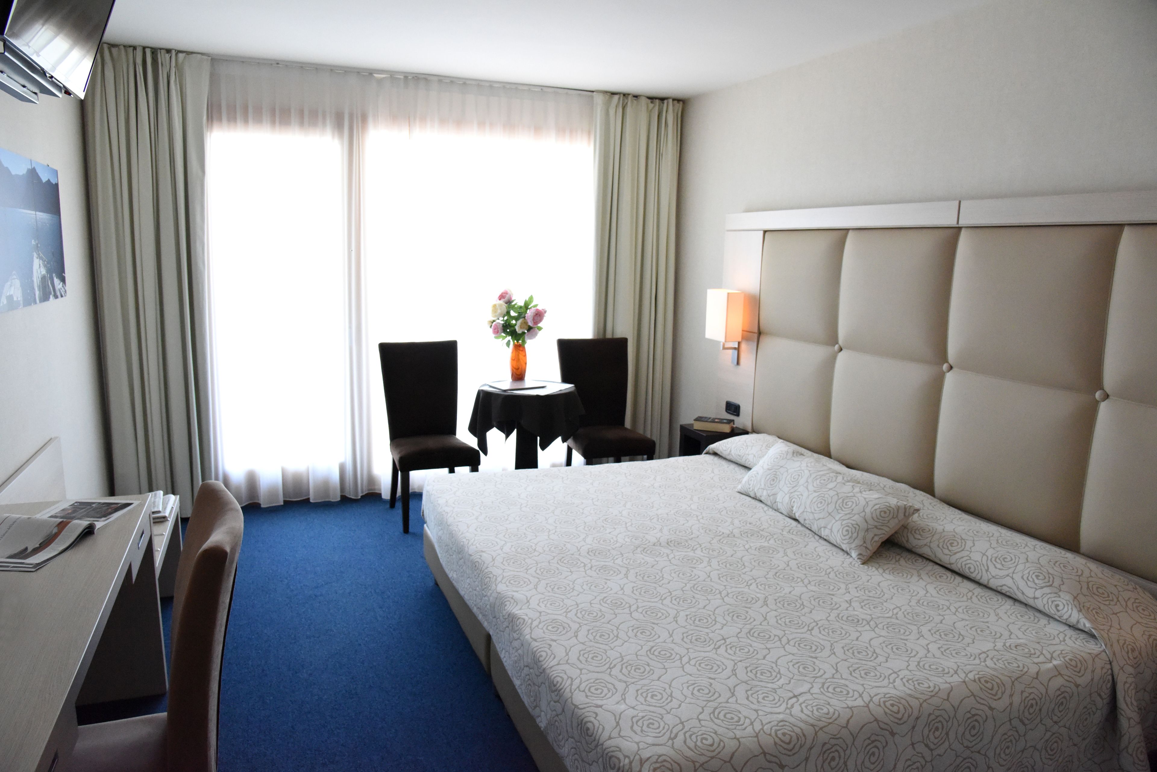 Ein Hotelzimmer mit einem großen Bett, einem Schreibtisch, einem Sessel, einem kleinen Tisch mit Blumen und Blick ins Freie durch das Fenster.