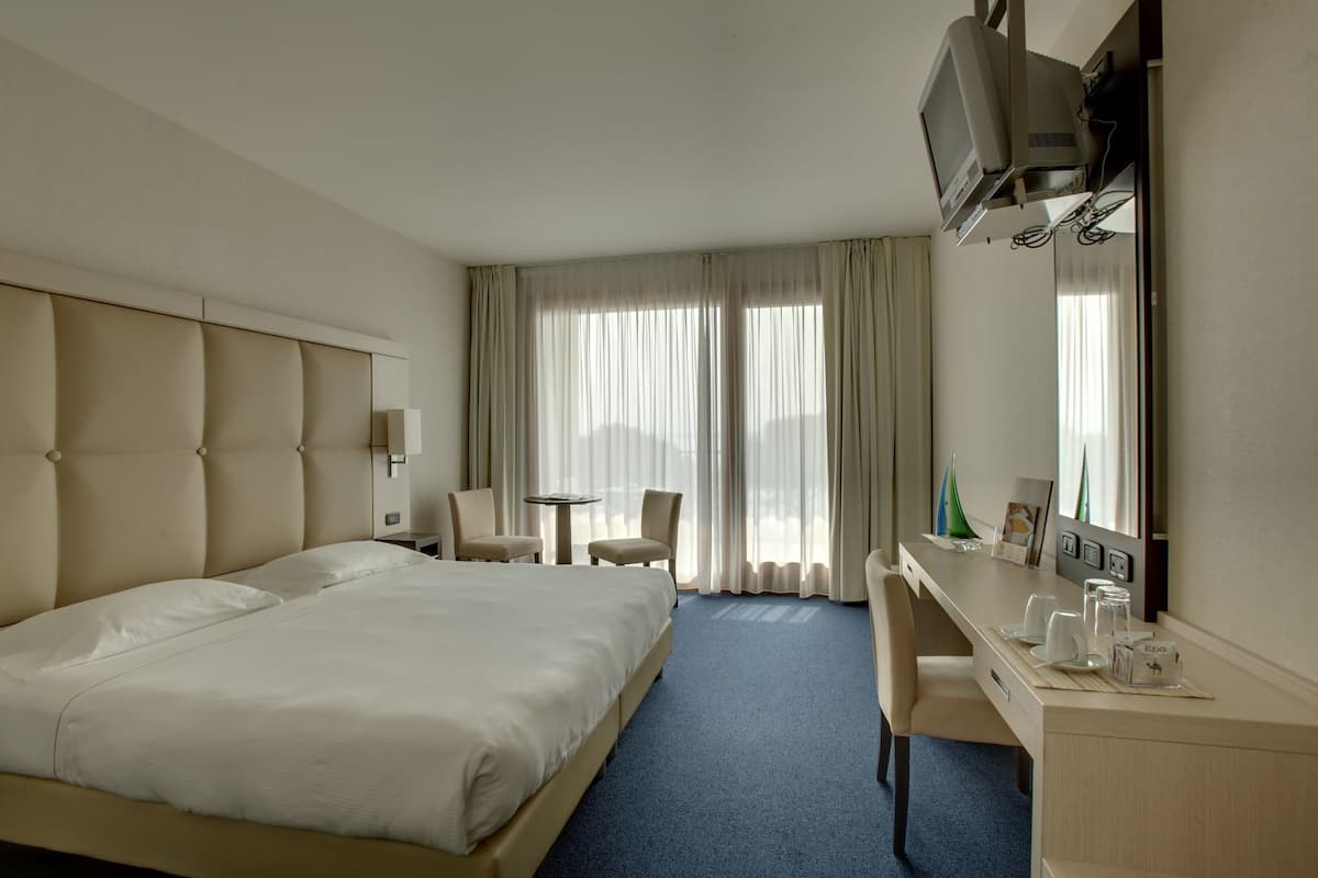 Ein Hotelzimmer mit einem großen Bett, einem Schreibtisch mit Stuhl, einem kleinen Tisch und einem an der Wand befestigten Fernseher.