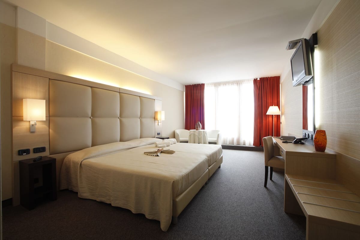 Ein Hotelzimmer mit einem großen Bett, einem Schreibtisch, einem Fernseher und einem Fenster mit Vorhängen.