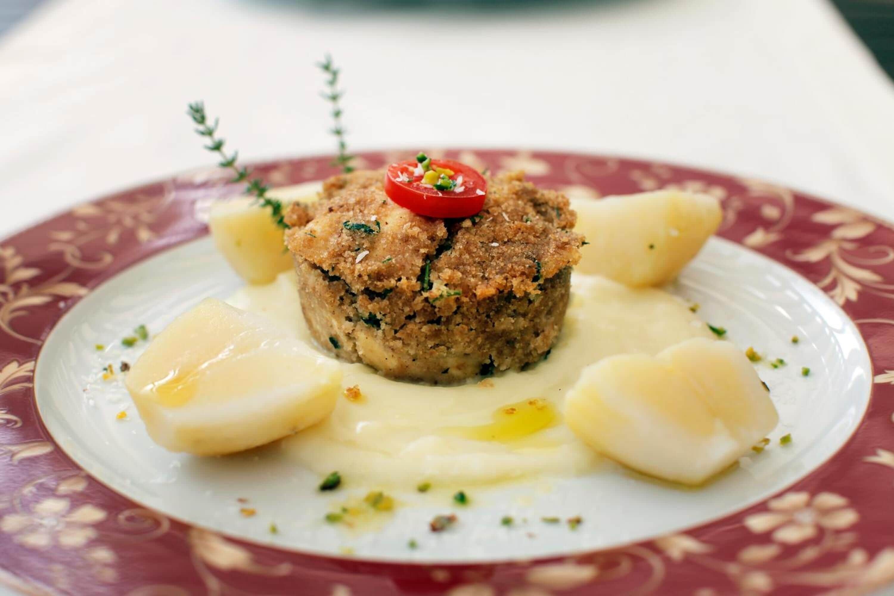 A dish with a breaded patty, a slice of tomato, and a creamy sauce on a patterned plate.