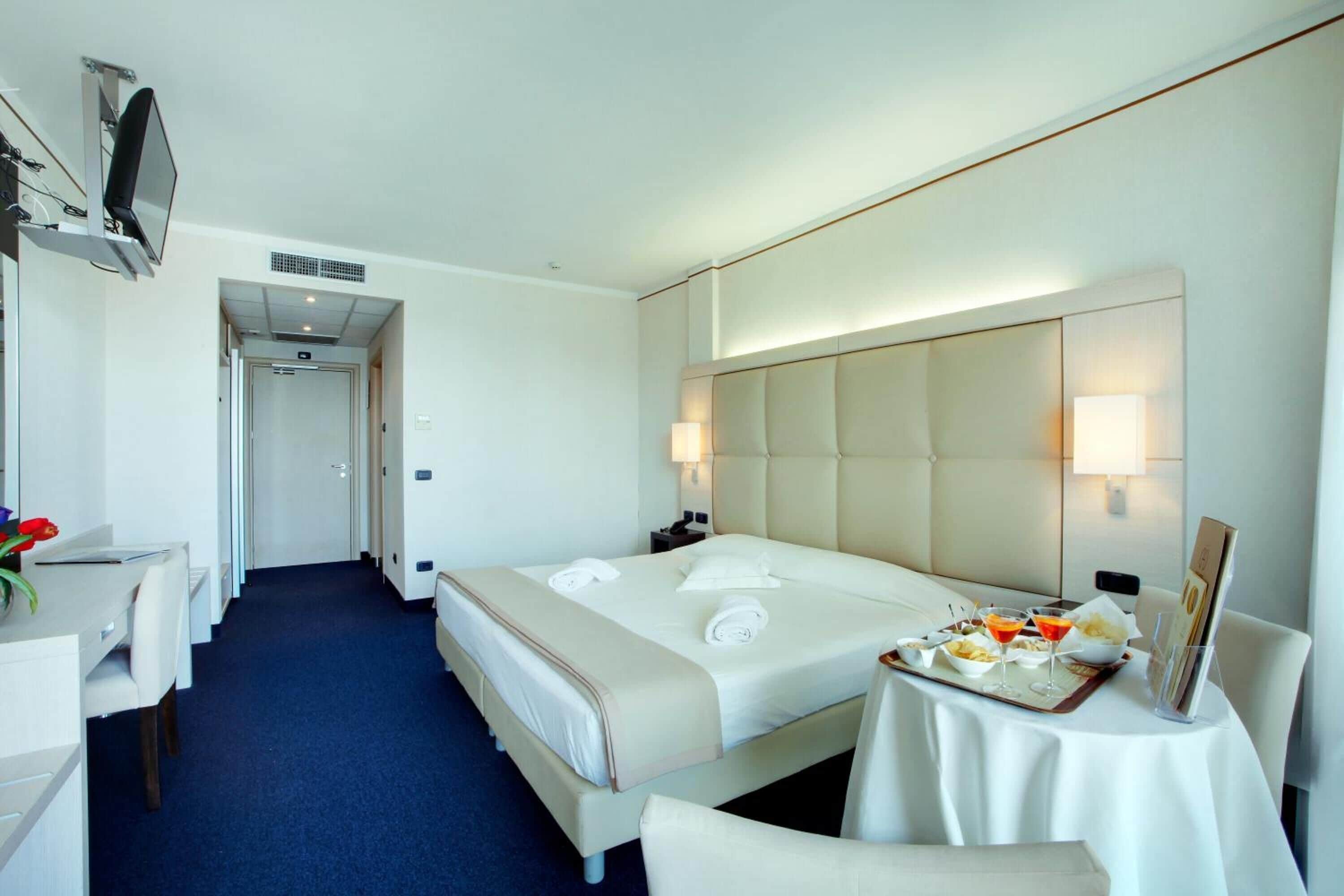 A hotel room with a bed, bedside table, desk, chair, and a small table with a tray of food and drinks.