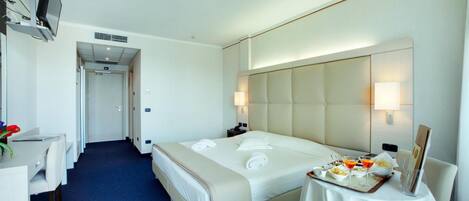 A hotel room with a bed, bedside table, desk, chair, and a small table with a tray of food and drinks.