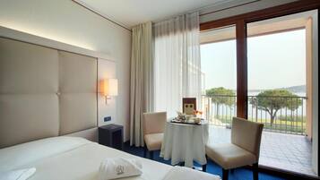 Ein Hotelzimmer mit einem großen Bett, einem kleinen Tisch mit Tablett, zwei Stühlen, einem Nachttisch, einer Lampe und einem Balkon mit Blick ins Freie.