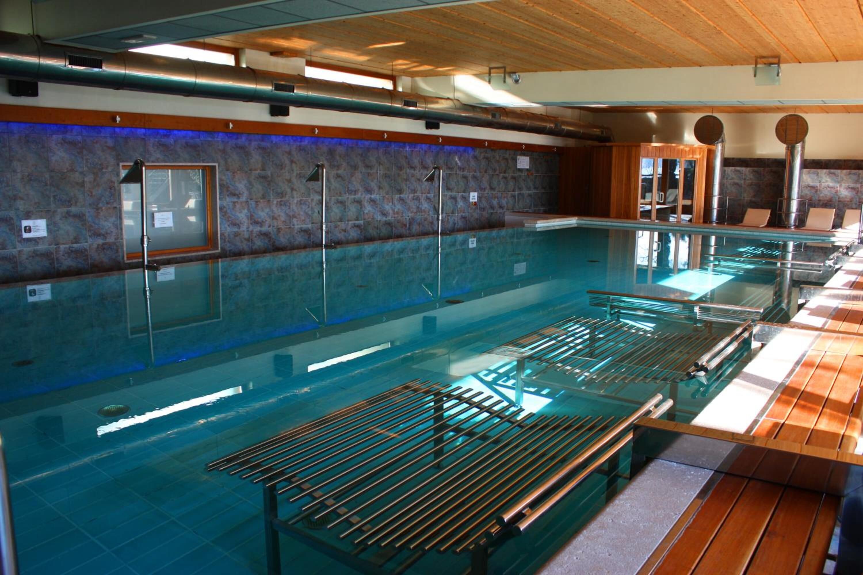 Una amplia piscina interior con varias pistas, una zona de terraza de madera y una pared de piedra con ventanal.