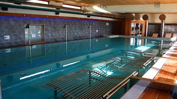 Una amplia piscina interior con varias pistas, una zona de terraza de madera y una pared de piedra con ventanal.