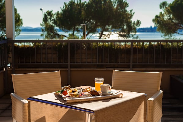 A balcony with wicker chairs, a table with breakfast, and a view of the water and trees.