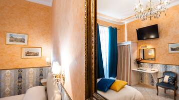 Quarto de hotel com um espelho grande, uma cama com almofadas azuis e amarelas, uma televisão na parede e um candelabro.