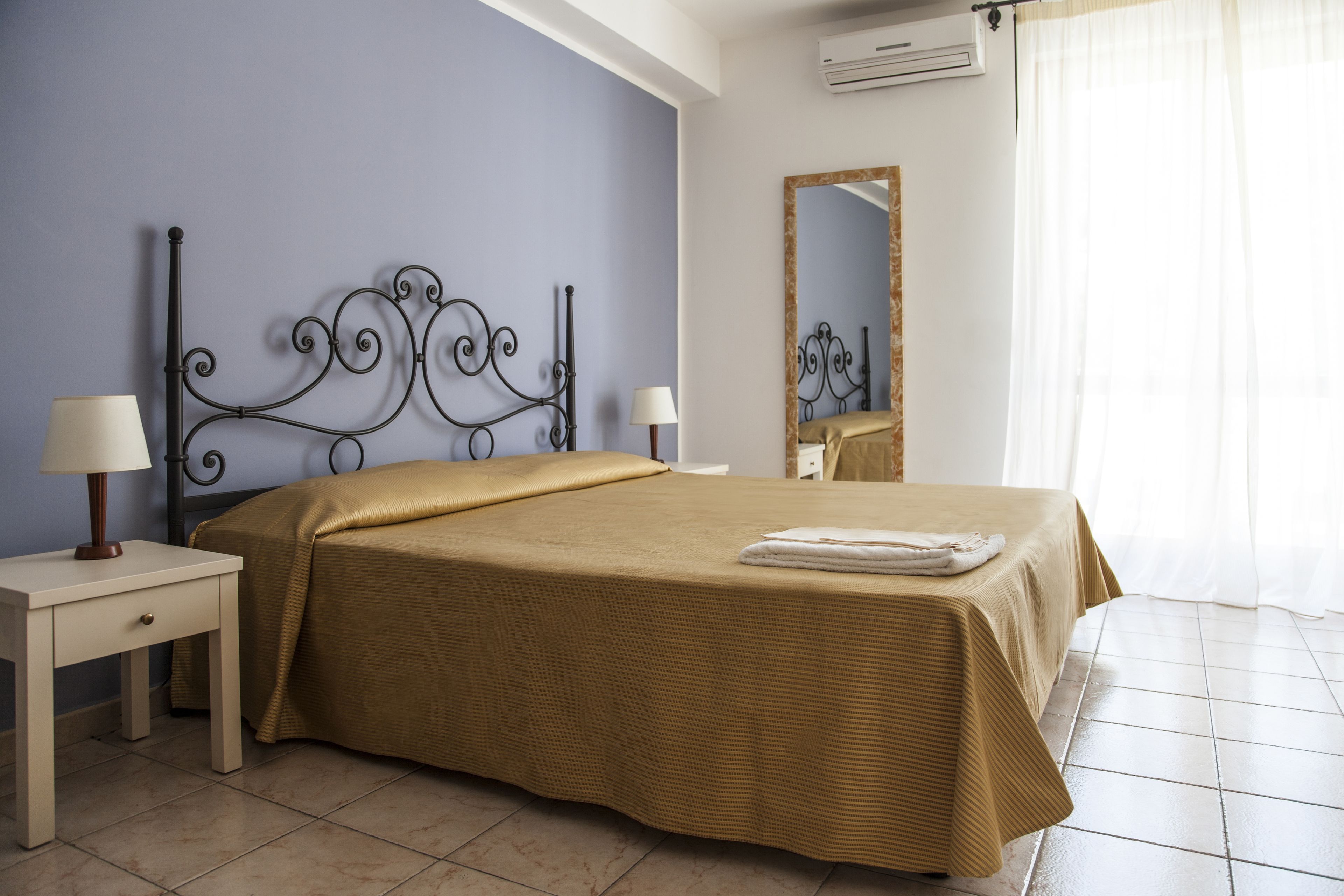 Une chambre à coucher avec un lit, deux tables de chevet, un miroir et une fenêtre avec des rideaux.