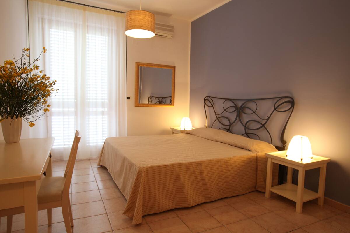 Un dormitorio con una cama, lámparas de noche, un espejo, una planta en maceta y una silla.
