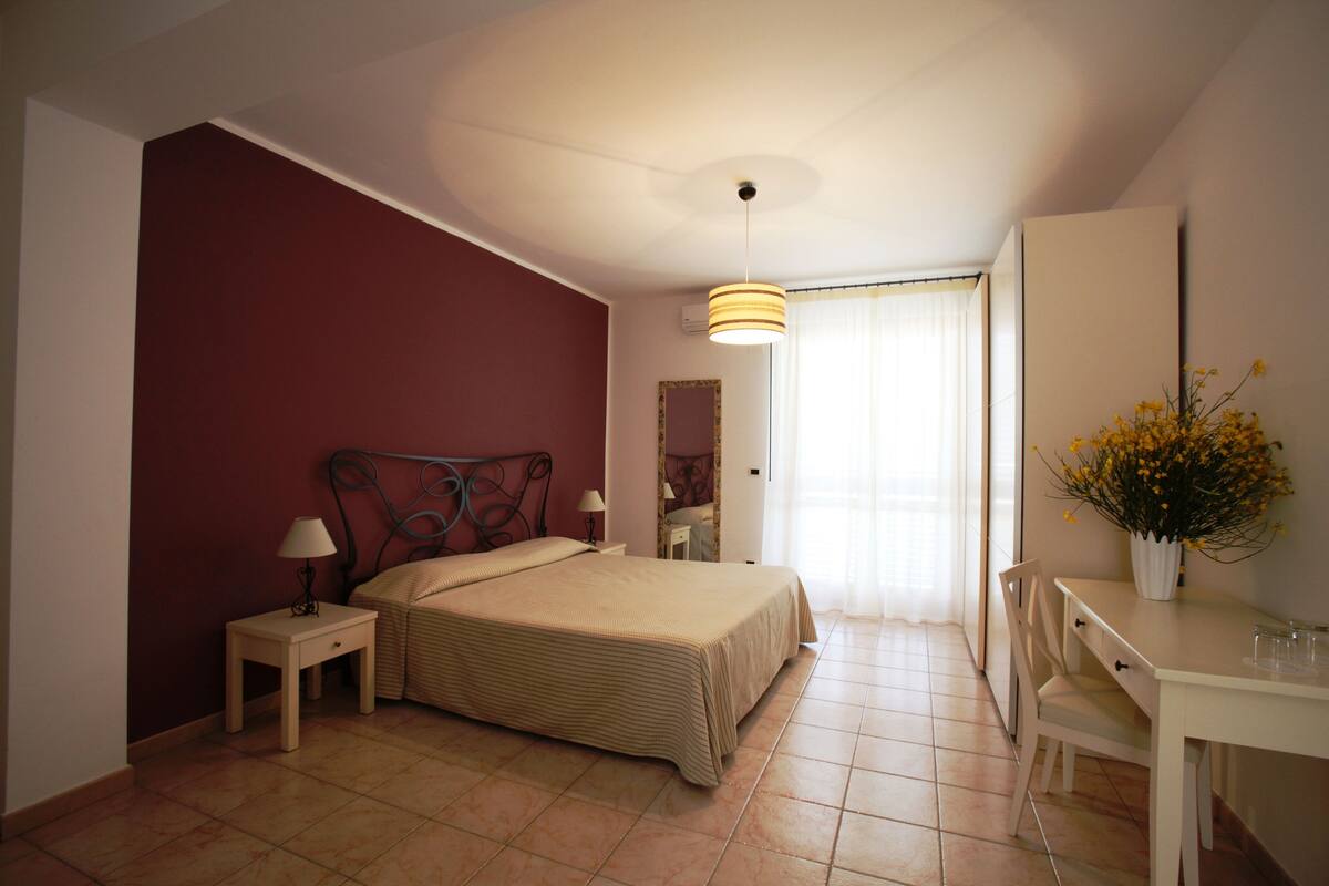 Un dormitorio con cama, mesita de noche, lámpara, silla, escritorio y un florero con flores.
