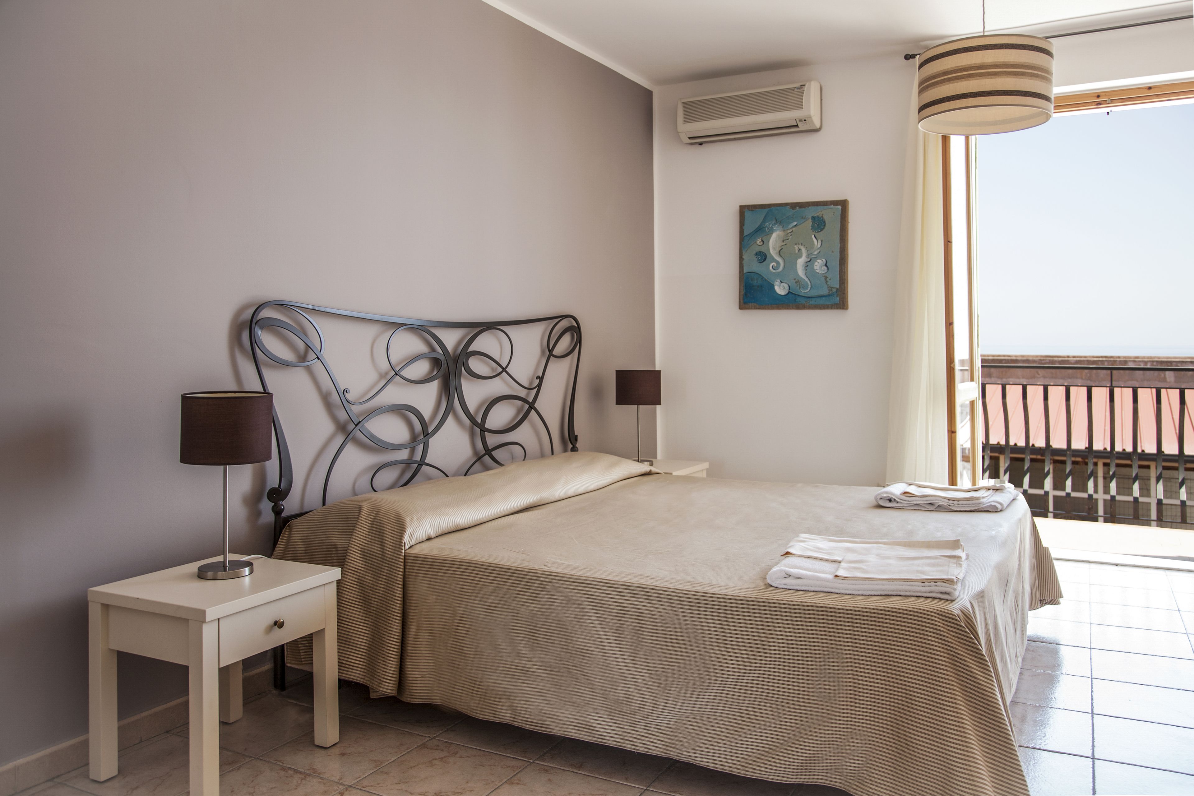 A bedroom with a bed, two bedside tables, a lamp, a painting, and a view of the outside.