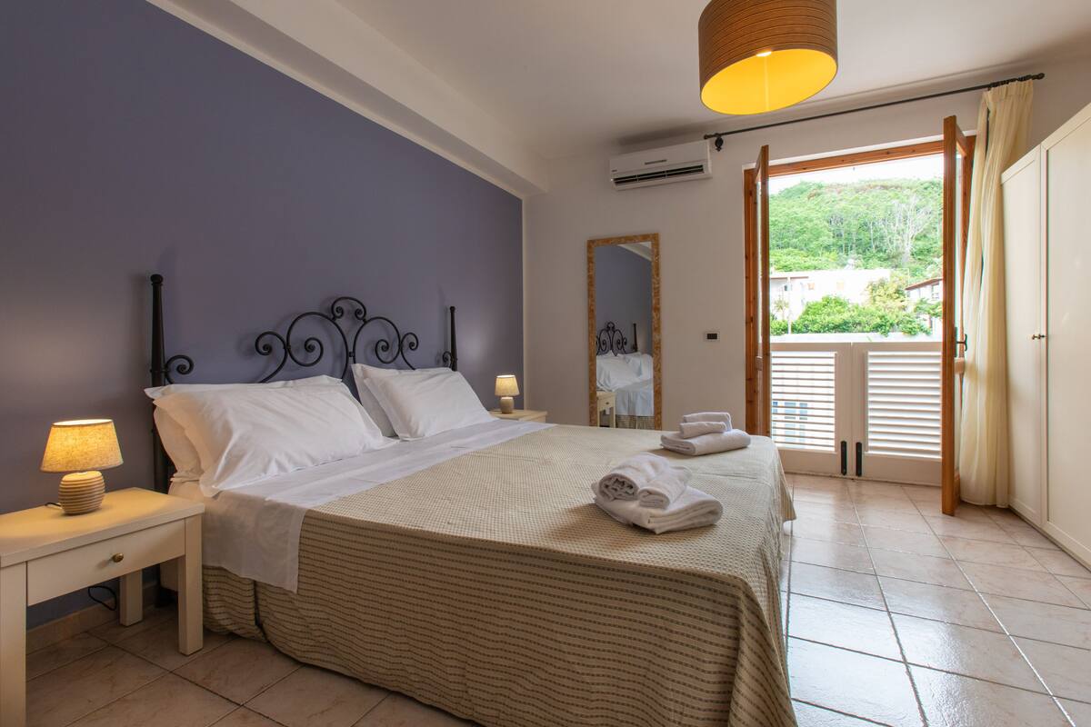Un dormitorio con cama, mesita de noche, lámpara, espejo y vistas al exterior.