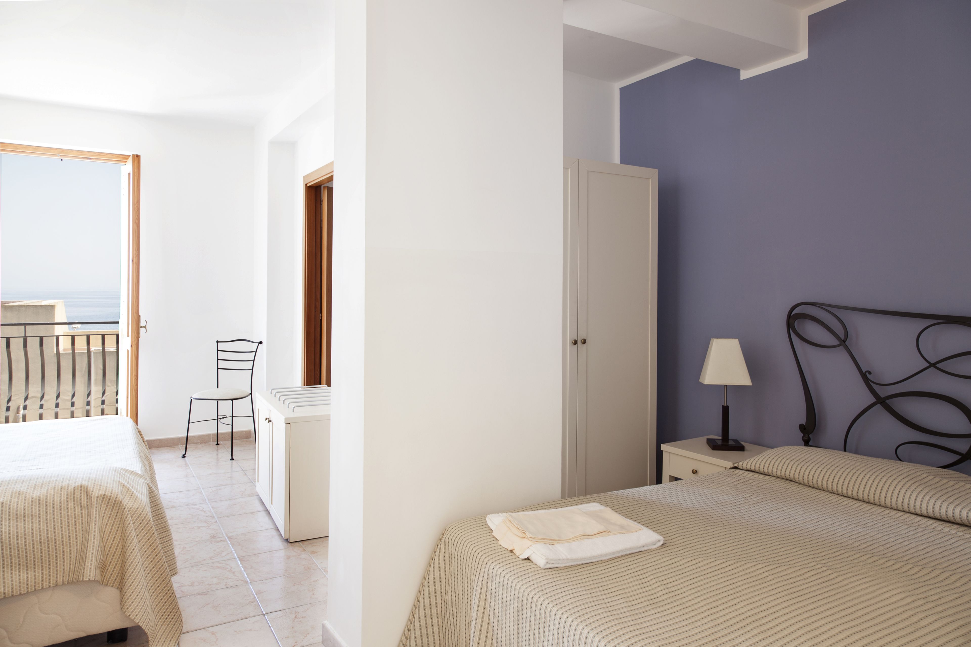 Une chambre avec deux lits, une armoire, une table de chevet avec une lampe, une chaise et une vue sur la mer par une porte ouverte.