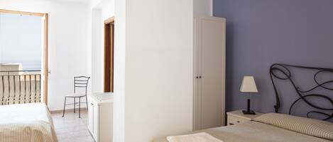 Une chambre avec deux lits, une armoire, une table de chevet avec une lampe, une chaise et une vue sur la mer par une porte ouverte.