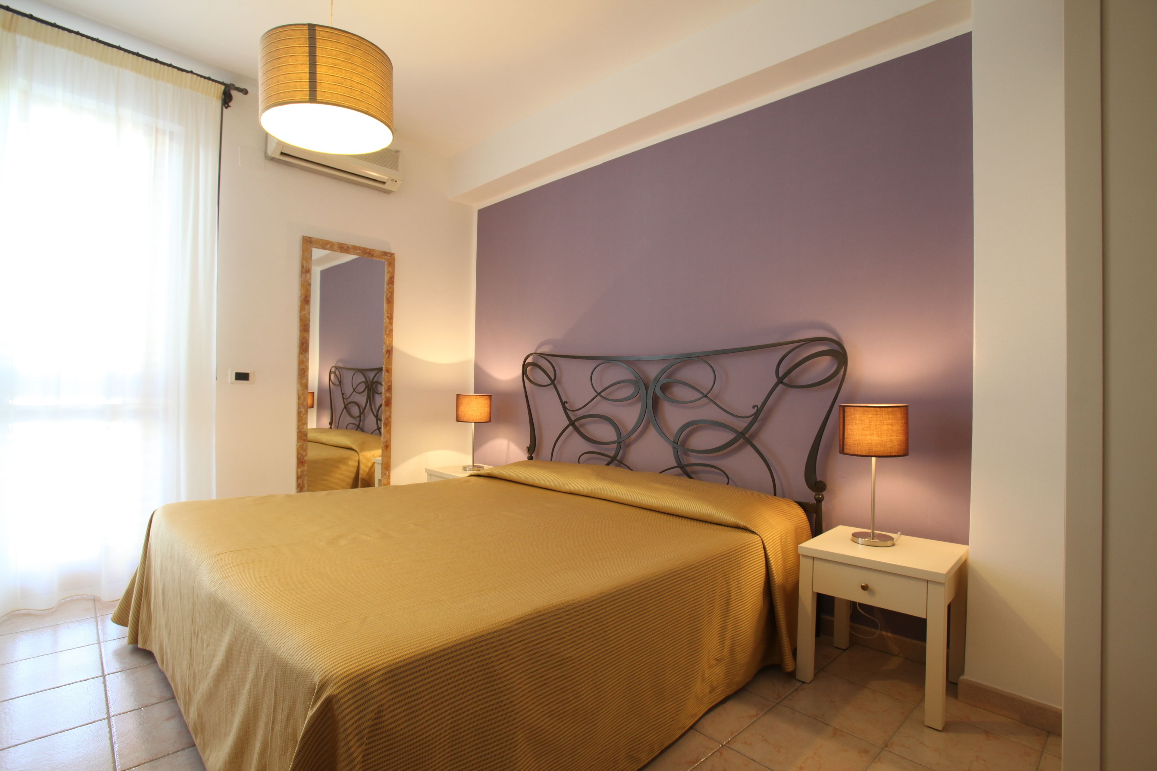 A neatly made bed with a gold bedspread, two bedside lamps, a mirror, and a purple wall.