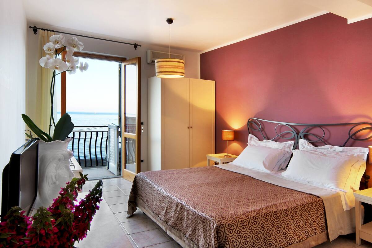 Habitación de hotel con cama, mesitas de noche, armario, balcón con vista al mar y una planta en maceta.
