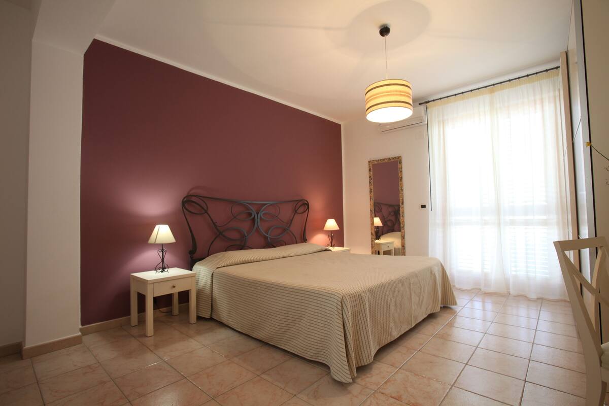 Un dormitorio con cama, lámparas de noche, mesita de noche, espejo y ventana con cortinas.