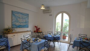 Une salle à manger avec des tables dressées pour quatre personnes, des nappes bleues et des chaises en métal. Un tableau orne le mur et une fenêtre offre une vue sur la verdure à l’extérieur.