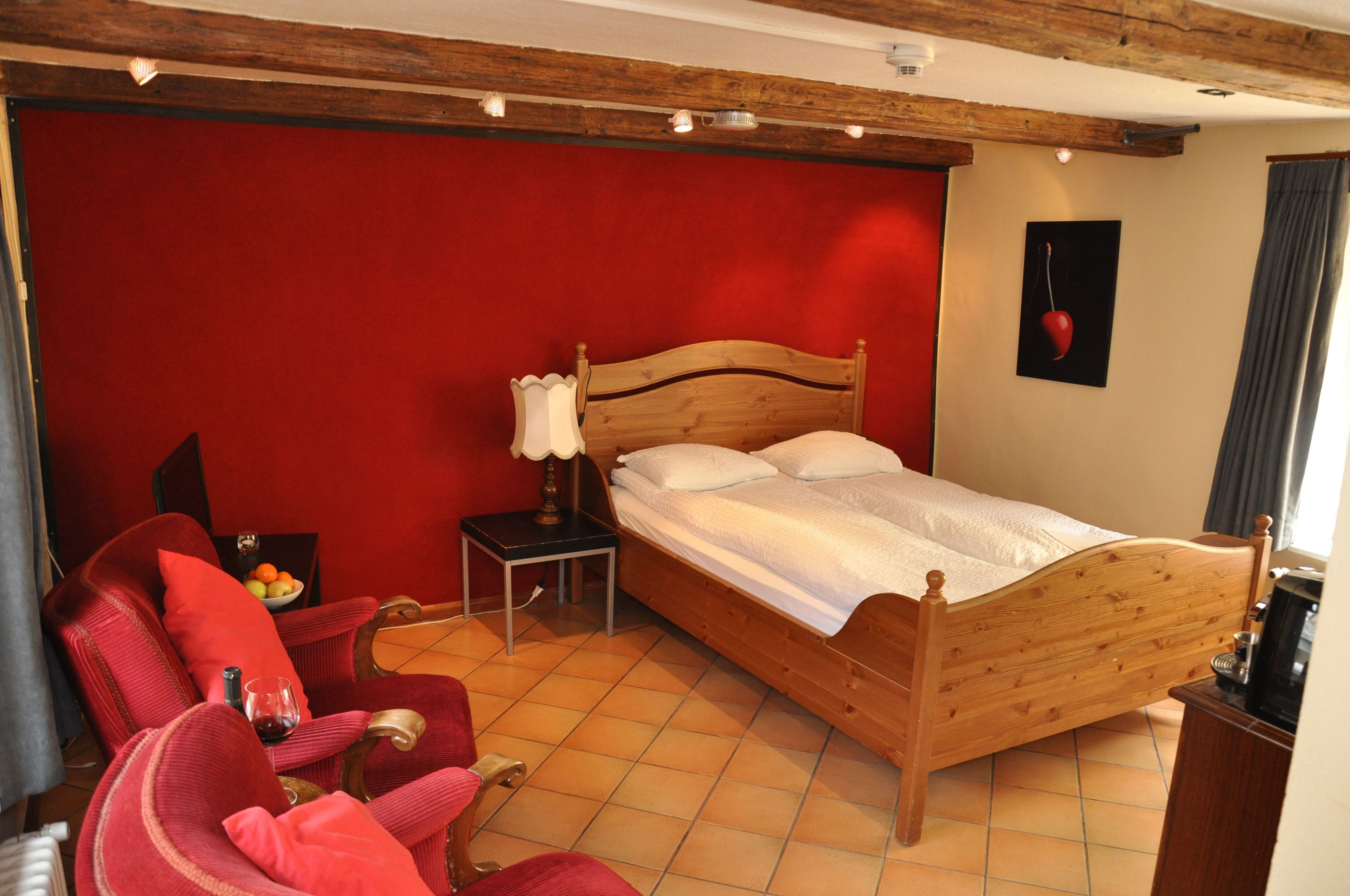 A bedroom with a wooden bed, a red wall, a small table with a lamp, and a chair.