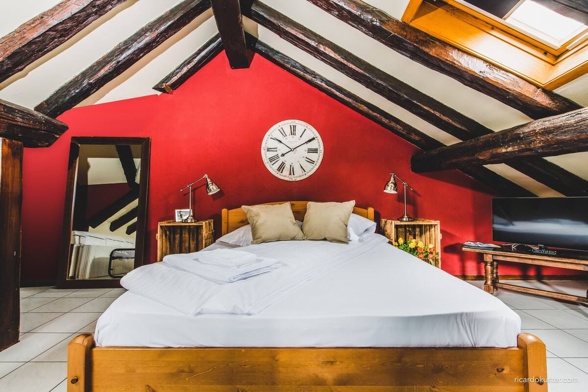 Un dormitorio con cama de madera, pared roja, reloj, televisor y ventana con vigas de madera.