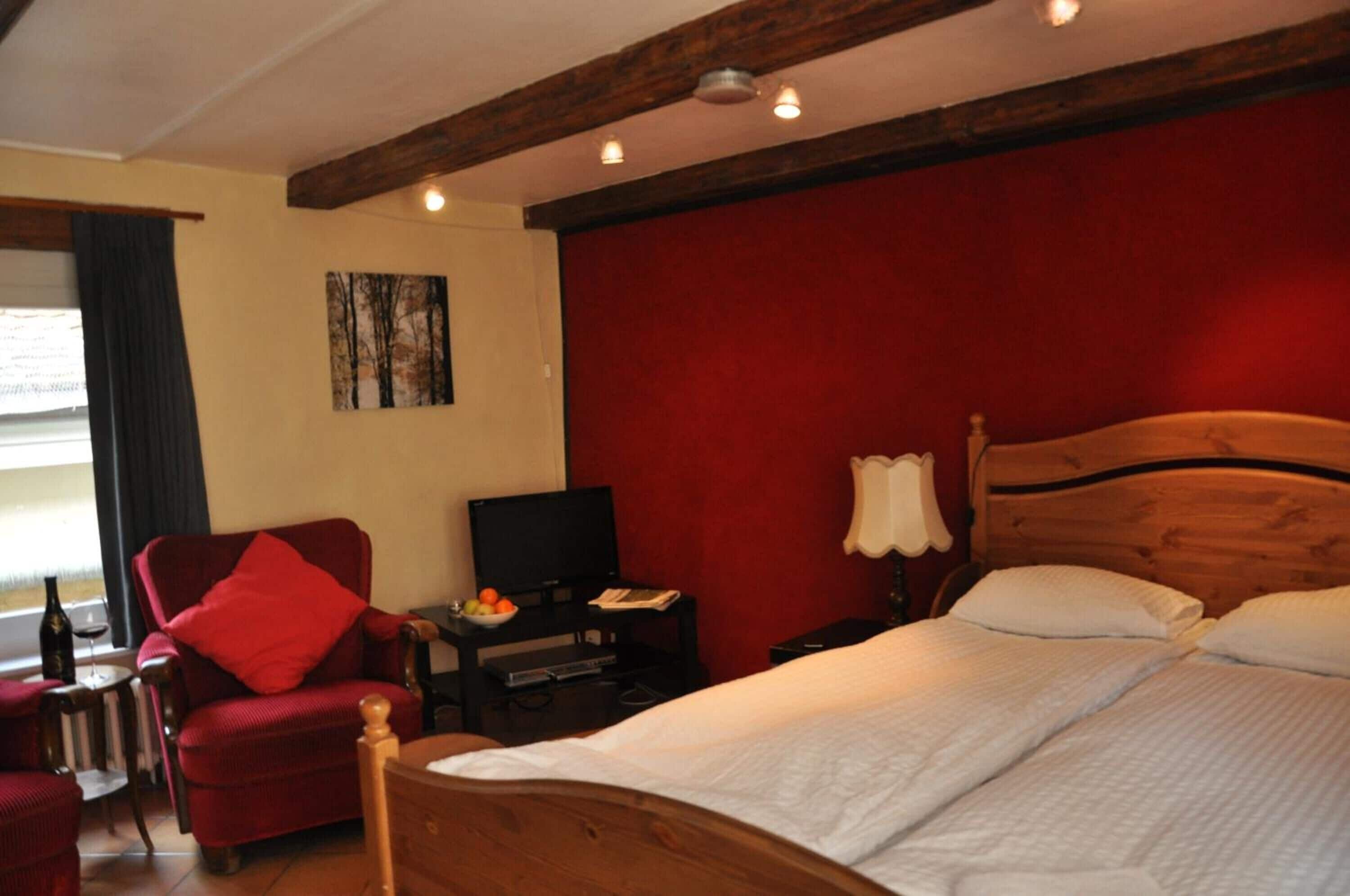Una habitación de hotel con una cama de madera, un sillón rojo, un televisor y una pared roja.