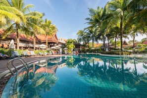 Une piscine entourée de chaises longues et de parasols, bordée de palmiers et de bâtiments aux toits rouges.