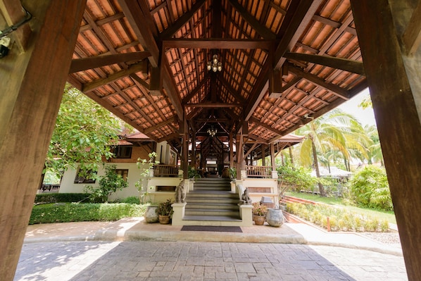 A covered staircase leading to a building with a wooden roof and columns, surrounded by greenery and palm trees.